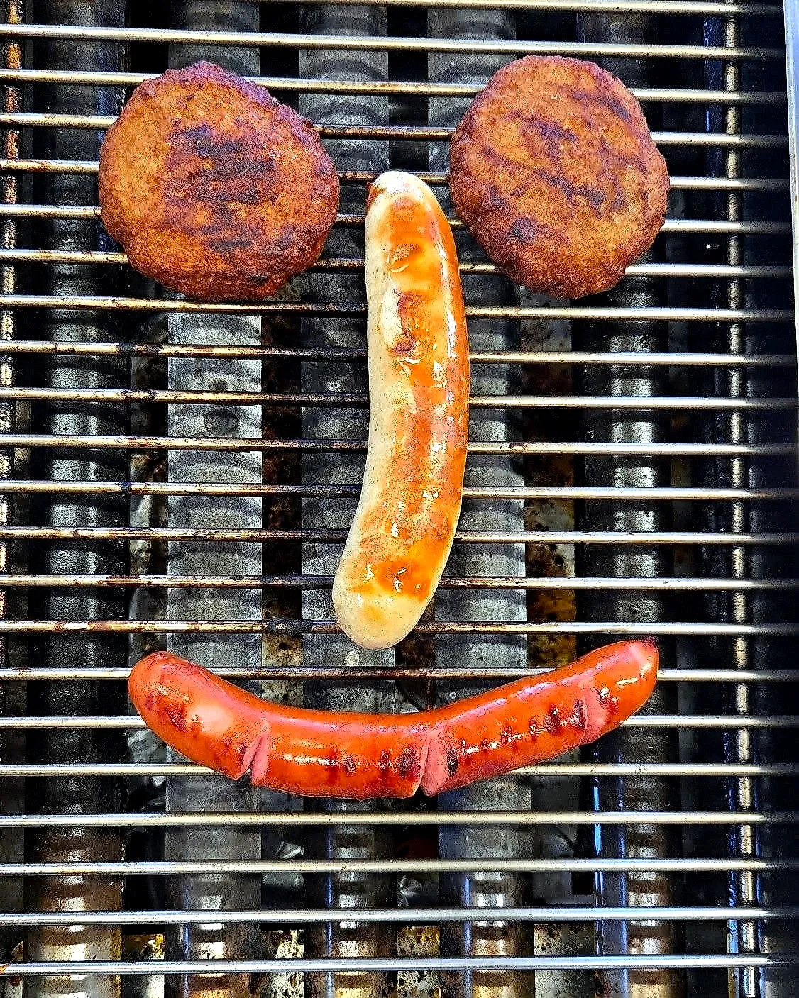 Ein Gesicht auf dem Grill: zwei Frikadellen als Augen, eine Bratwurst als Nase, eine Krakauer als Lächeln